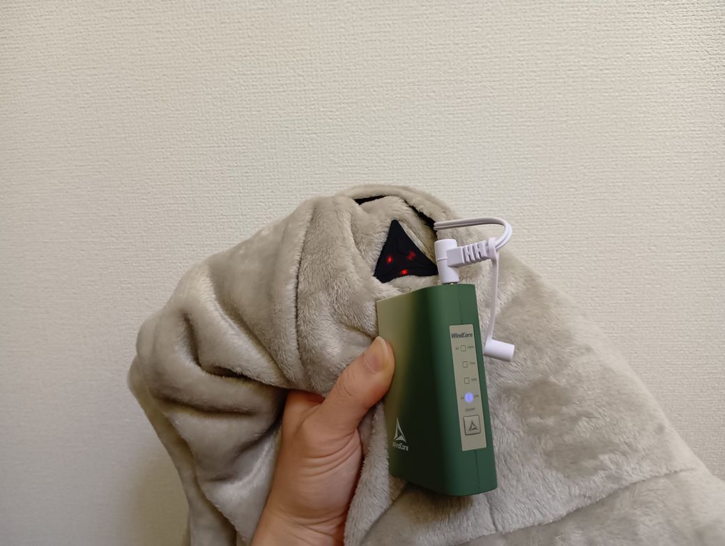 着るコタツに付いてる白色の有線を充電器に接続。電源を入れると、黒色の三角マーク（温度調整スイッチ）が赤色に点滅→服をポカポカにしてくれる（写真：筆者撮影）