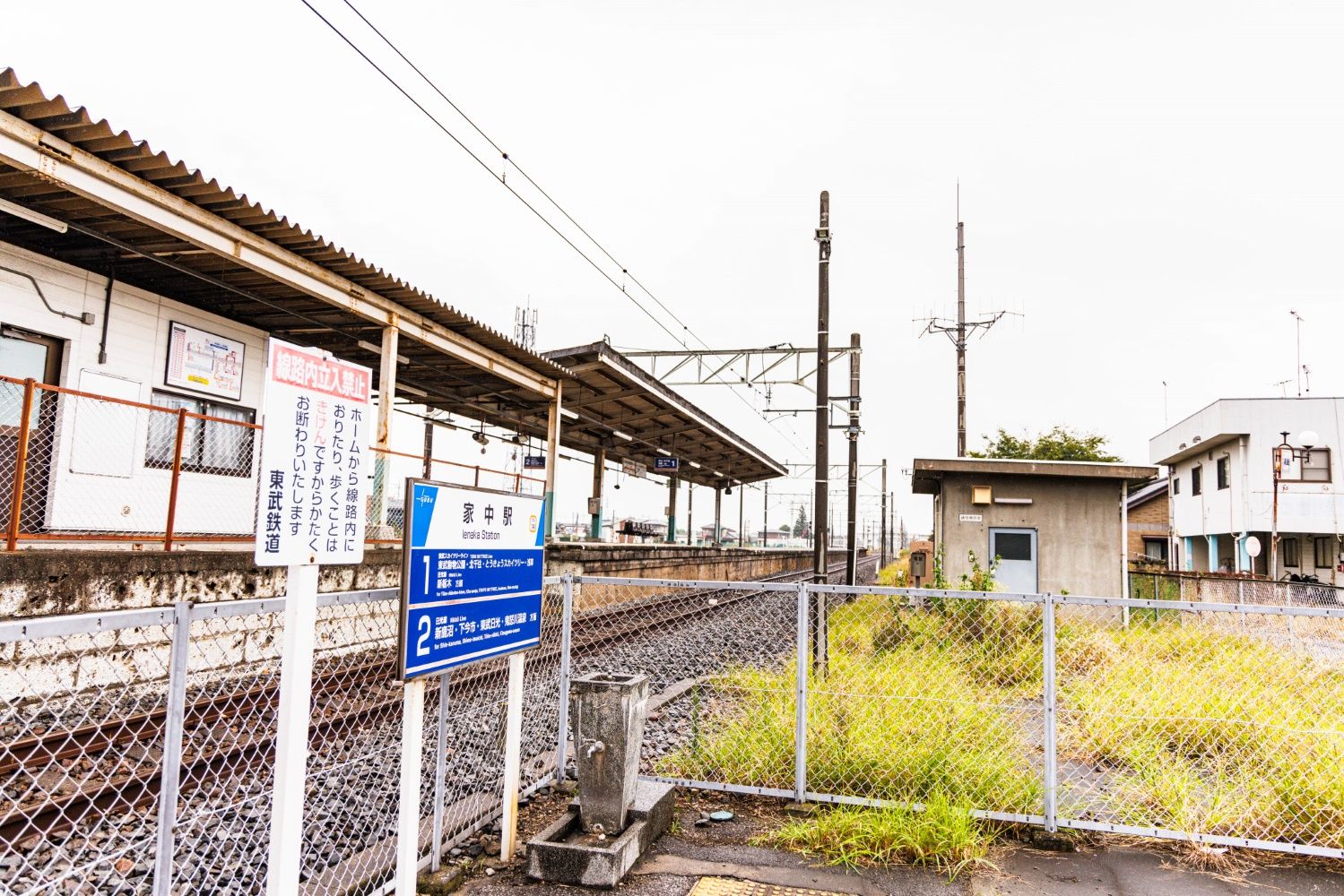 ホームにありそうな水飲み場が家中駅東口の駅名板の脇にポツン。柵の向こうに線路が通る（撮影：鼠入昌史）